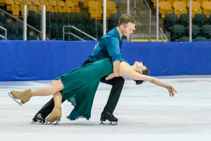 A woman lays back as her partner supports her behind her shoulders with his hands and his knee. They're low to the ice. 